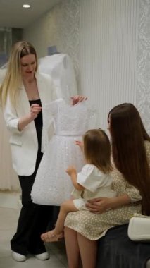 Gülümseyen kadın, şık gelinlik salonunda annesinin kucağında oturan küçük kıza parlak elbiseler gösteriyor..