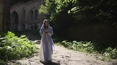 Uzun beyaz bir bornoz giymiş ve açık mavi bir pelerin giymiş genç bir kız, yemyeşil yapraklarla çevrili, güneşli, kirli bir yolda yürüyor..