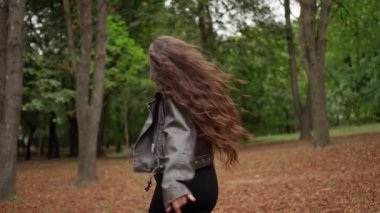 Görüntü, parkta dönen neşeli hamile bir kadını gösteriyor. Şık, koyu renk deri bir ceket ve siyah bir elbise giyiyor..