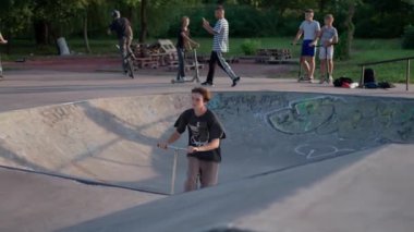 Genç çocuk arka planda arkadaşları ve diğer motosikletçilerle birlikte kaykay parkının beton kasesinde ustalıkla scooter manevrası yapıyor..