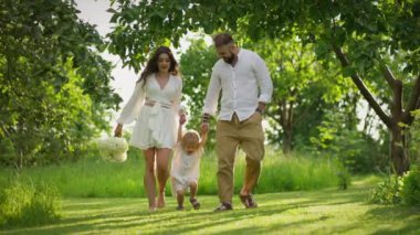 Neşeli ebeveynler, yeşil ve ağaçlarla çevrili güneşli parkta yürürken küçük çocuklarının ellerini tutuyorlar..