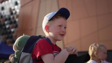 Beyzbol şapkalı ve sırt çantalı küçük çocuk neşeli bir şekilde dışarıdaki olayları izliyor, merak ve macera duygusunu somutlaştırıyor..