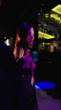 Kendinden emin bir kadın, siyah cüretkar bir elbise içinde poz verir. Saçları, modern bir salondaki dinamik sarı ve mor ışıkla aydınlatılmış canlı ve şık bir atmosferden zevk alırken süzülür..