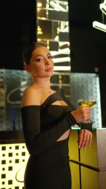 Elinde şampanya kadehi tutan şık siyah kıyafetli, modern, aydınlık bir arka planda metalik ve geometrik tasarımlar içeren dramatik ışıklandırma altında poz veren şık bir kadın..