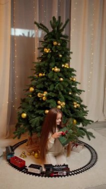 Konforlu krema kıyafetli küçük kız, Noel ağacının yanında altın süslemelerle süslenmiş, güzel bir hediye tutuyor, oyuncak trenle çevrili, festival havasında..