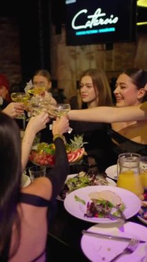Canlı bir grup insan, renkli meyve, salata ve içeceklerle dolu güzel dekore edilmiş yemek masasında kadeh kaldırırken şampanya kadehlerini tokuşturur..
