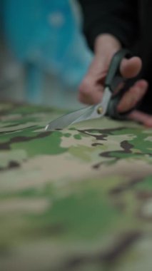 Masadaki yeşil ve kahverengi kamuflaj desenli kumaşı kesmek için makasları dikkatlice kullanın. Tekstil işindeki ustalığı ve hassasiyeti vurgulayın..