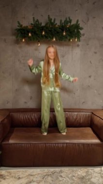 Uzun sarı saçlı, parlak yeşil pijama giyen neşeli bir çocuk yeşil ve sıcak ampullerle süslenmiş rahat bir iç dekorasyonda kahverengi deri bir koltuğa atlıyor..