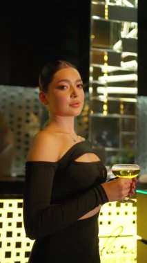 Dar siyah elbiseli zarif kadın modern cazibe ortamında sıcak ortam ışıkları altında kokteyl bardağı tutarken zarif poz veriyor..