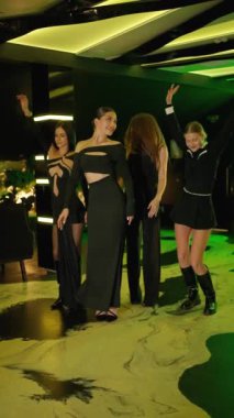 Gece kulübündeki dört şık kadın grubu dans ediyor ve gülümsüyor, zarif siyah gece elbiseleri ve ayakkabıları giyiyorlar, hareketli gece atmosferinin tadını çıkarıyorlar..