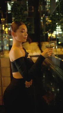 Sofistike siyah elbiseli şık genç bir kadın, elinde kokteyl bardağıyla, loş ışıklı modern bir iç mekanda güvenle duruyor..