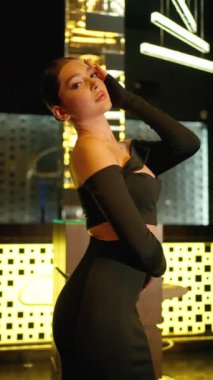 Gösterişli siyah bir elbise giymiş şık bir genç kadın, bir eli modern bir ortamda güvenli bir şekilde poz veriyor, parlak geometrik aydınlatmalar ve parlak bir arka planla vurgulanıyor..