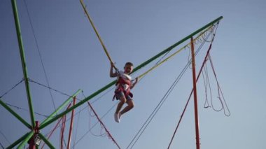 Koşumlu genç bir çocuk açık mavi bir gökyüzünün altında bungee trambolinde zıplıyor..