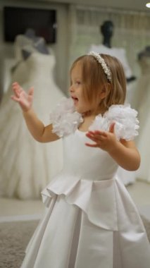 Omuzlarında çiçek desenleri olan beyaz elbiseli küçük bir kız ve gelinliklerle çevrili gelinlik butiklerinde duran inci saç bandı..