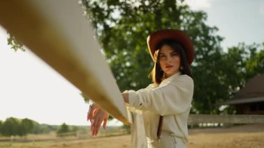 Geniş kenarlı bir şapka ve beyaz bir kıyafet giyen şık bir kadın ağaçlarla ve açık arazilerle güneşli bir kırsalda ahşap bir çite yaslanıyor..