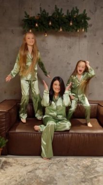 Yeşil ipek pijama giyen bir kadın ve iki çocuk, yeşil ve dekoratif ışıkların altında kahverengi bir deri koltukta gülüp oynuyorlar..