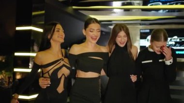 Zarif siyah kıyafetler giymiş dört genç kadın modern, loş bir mekanda bir araya gelip gülüyorlar ve birbirlerine sarılıyorlar. Mutluluk ve dostluk yayıyorlar..