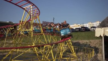 Bir grup insan, parlak güneşli bir günde binaların yakınında canlı, sarı ve kırmızı bir lunapark trenine biniyor..