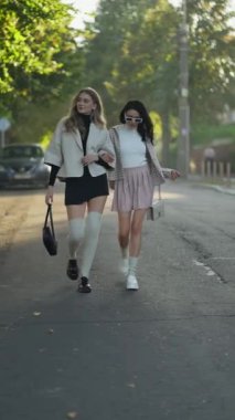 Şık kıyafetler giyen iki şık kadın gün ışığında ağaçlarla kaplı sessiz bir sokakta birlikte yürüyorlar..