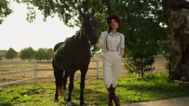 Şapkalı ve çizmeli şık bir kadın kırsal kesimde siyah bir atın yanında duruyor..