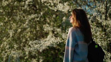 Güneş gözlüğü ve pastel süveter giyen bir kadın ağaçların yanında beyaz çiçeklerle açan güneş ışığında duruyor..