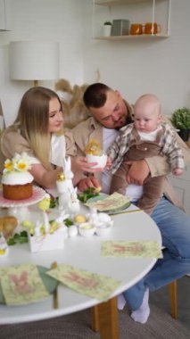 Bebekli bir aile, Paskalya temalı süslemeler ve ikramlar ile süslenmiş bir şenlik masasında oturur..