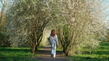 Sıradan giyinmiş bir kadın, çiçek açan ağaçlar ve yemyeşil çiçeklerle dolu sakin bir yolda yürüyor..