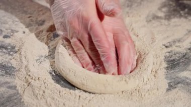Dağınık un ile kaplı karanlık bir yüzey üzerinde çiğ pizza hamuru şekillendiren eldivenli eldiven..