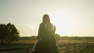 Gün batımında bir çiftlikte siyah ata binen bir kadın, manzaralı bir kırsal arka planı var..