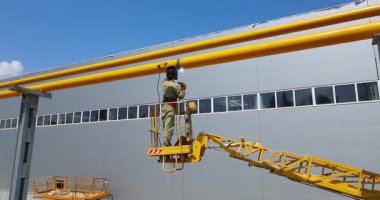 Hava asansöründe koruyucu giysili bir işçi büyük bir endüstriyel binanın dışında sarı borular bulundurur..