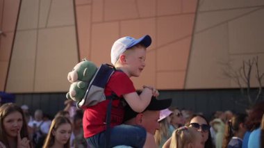 Kırmızı tişörtlü ve şapkalı gülümseyen bir çocuk açık hava etkinliğinde babasının omuzlarına oturur, oyuncak bir sırt çantasıyla.