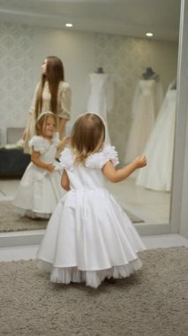 Beyaz elbiseli, çiçek desenli genç bir kız aynada kendine bakarken arka planda elbisesiyle duran bir kadın....