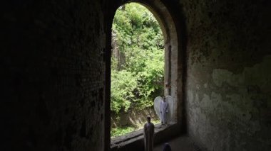 Biri melek kanatlı, beyaz cüppeli üç kişi yeşillik yakınlarındaki çürümüş tuğla bir yapıda duruyor..