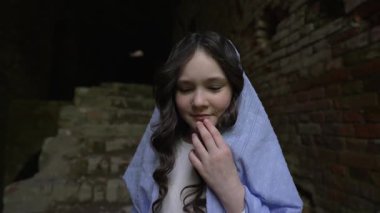 Uzun kıvırcık saçlı, açık mavi atkı takan ve beyaz elbiseli genç bir kız loş bir alanda eski taş merdivenlerin yanında duruyor..