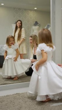 Küçük kız aynanın önünde beyaz bir çiçekçi kız elbisesi deniyor, yetişkinlerin yardımıyla, düğün için hazırlanıyor, gülümsüyor ve neşe saçıyor..