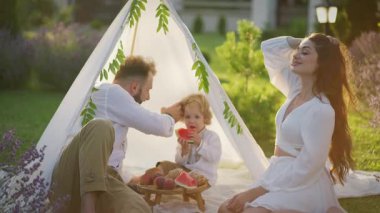 Ebeveynler ve küçük oğulları, akşam ışığında lavanta tarlalarıyla çevrili çadırın yanında karpuzun tadını çıkarırlar..