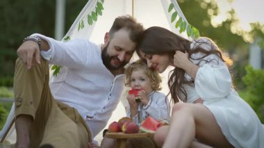 Doğada mutlu aile pikniği, genç ebeveynler ve çocukları karpuz ve meyvelerin tadını çıkarıyorlar, sevgi ve gülümsemeyle rahat beyaz bir çadırda dinleniyorlar..