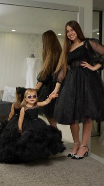 Güneş gözlüklü genç bir kızın neşeli tavırları, her ikisi de parlak siyah giysiler giyen bir kadının zarafetiyle eşdeğerdir..