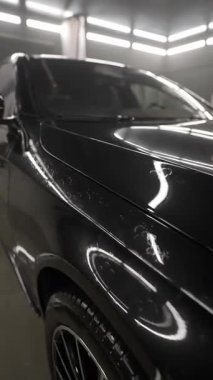 Su lekeleri olan siyah arabanın yakın çekimi, atölyedeki ışıkları yansıtıyor. Islak yüzey ve parlak boya, profesyonel oto temizleme ve cilalama ortamı.