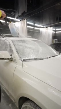 Sarı takım elbiseli bir işçi, araba yıkama bölümünde köpük spreyli beyaz araba yıkıyor, temizlik işlemi gösteriliyor..