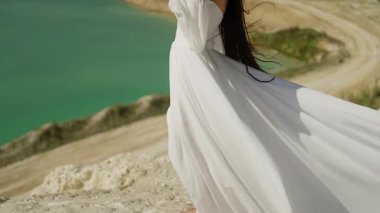 Beyaz elbiseli bir kadın turkuaz bir gölün yanında duruyor. Güneşli bir günde, deniz kenarında uzun elbiseli zarif bir kadın figürünün doğal bir manzaranın arka planına karşı yakın plan fotoğrafı..