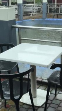 Boş kafeterya masası, yağmurdan sırılsıklam sandalyeler, siyah plastik koltuklar ve beyaz masa üstünde oluşan su birikintileri. Boş havuz ve teras alanının yanında yağış sırasında karamsar kentsel an görüntülendi.