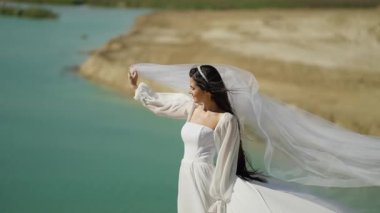 Gelin denizin kenarında duruyor, rüzgar neşeyle örtüsünü kaldırıyor dingin bir gölün arka planına.