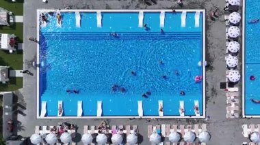 Yüzücülerle ve şemsiyeli güneşliklerle dolu büyük bir yaz yüzme havuzunun tepeden aşağı görüntüsü..