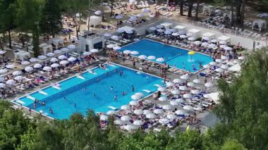 Kalabalık bir tatil köyü, birden fazla havuz, güneş pansiyonları ve ormanla çevrili beyaz şemsiyeler, canlı yaz atmosferini ve dinlenme rahatlığını yakalıyor..