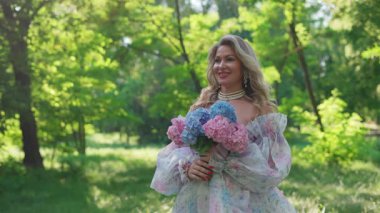 Elegant woman with blonde hair holding a bouquet of pink and blue hydrangeas, wearing a floral dress and pearls in a green summer park.