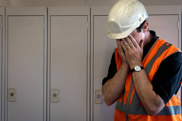 A male employee with his head in his hands in despair struggling to cope at work in an industrial or construction environment in a mental health concept