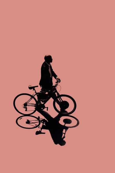 Silhouette of a male businessman walking alongside and pushing a bicycle with a plain pink background and copy space