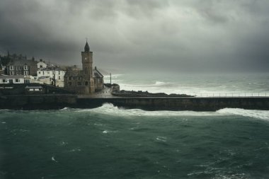 Dark storm clouds with gale force winds and waves crashing against the harbour wall and church in Porthleven on the Lizard peninsula in Cornwall clipart