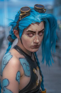 Savaşa hazır ve Jinx gibi giyinmiş mavi saçlı güzel bir kız kameraya özgüvenle bakıyor.